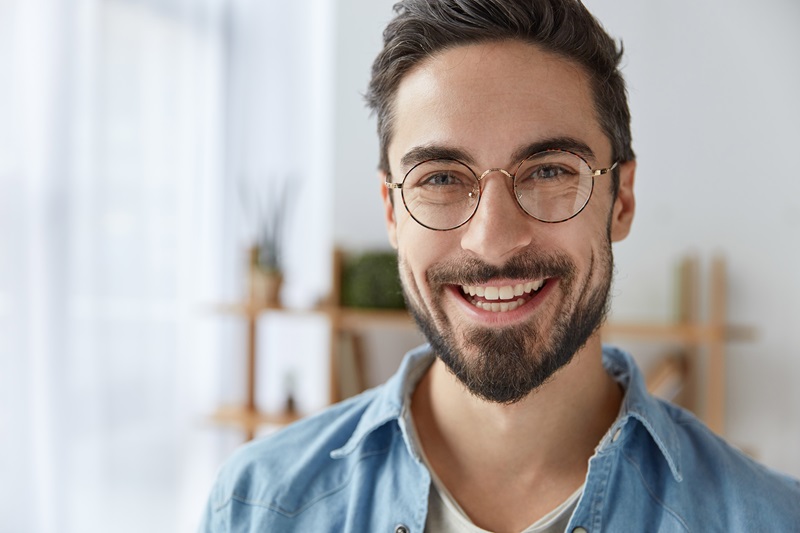 Ein junger Mann mit Brille und Vollbart steht in einer Wohnung und lacht in die Kamera.