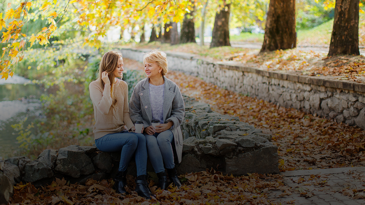 Eine jüngere Frau sitzt mit einer älteren Frau auf einer niedrigen Steinmauer. Sie trinken Kaffee aus einem Becher.