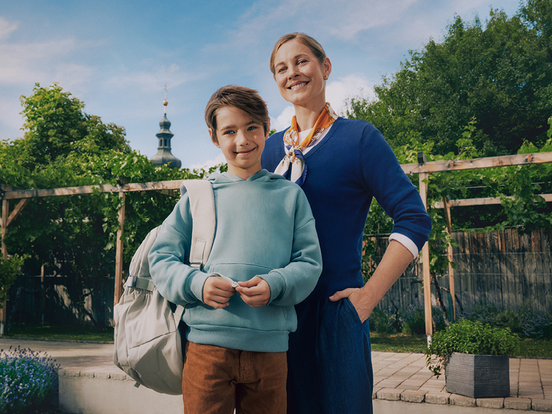 Eine Mutter und ihr Sohn stehen hintereinander. Die Mutter trägt einen dunkelblauen Pullover und ein Tuch, der Sohn einen hellblauen Hoodie und eine Schultasche über der rechten Schulter.