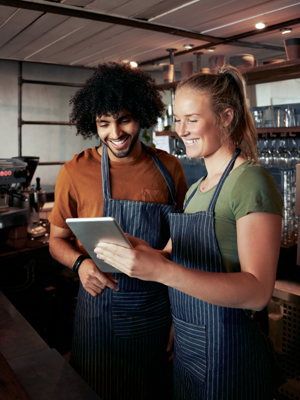 Ein Mann und eine Frau stehen in ihrem Kaffeehaus und blicken lächelnd auf ein Tablet.