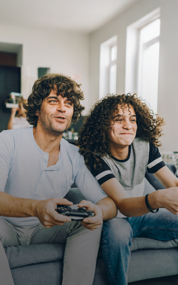 Ein Vater und sein Sohn sitzen auf einer Couch und spielen Videospiele mit Controller in der Hand.